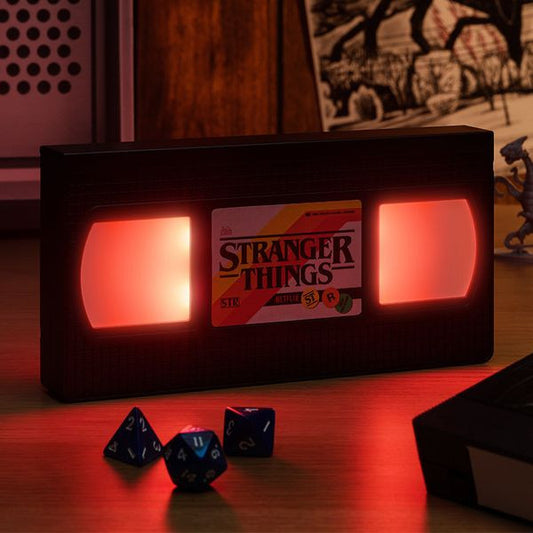 Stranger Things VHS tape lamp with glowing red accents, surrounded by colorful dice and retro decor elements.