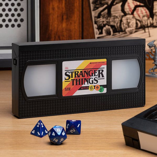 Stranger Things VHS-inspired box on a wooden table, featuring colorful branding and nearby blue dice.
